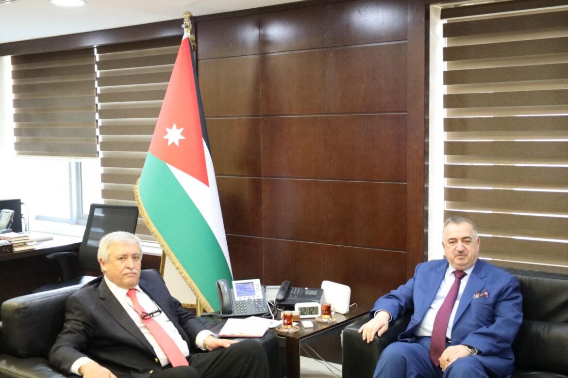 Two men in suits sit on black leather couches in a formal office with a Jordan flag between them.